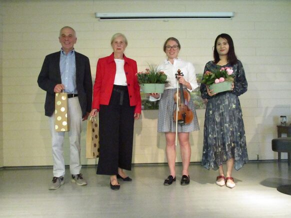 Zu sehen sind vier Personen. Links ein Mann, rechts daneben drei Frauen. Sie halten Geschenktüten, Pflanzen und eine Violine in der Hand